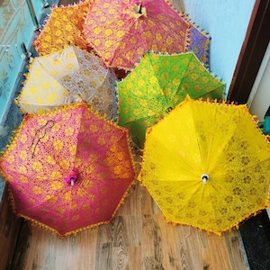 May include: A collection of colorful, decorative umbrellas with intricate floral patterns. The umbrellas are in shades of pink, yellow, green, and orange, with gold accents and tassel trim. The umbrellas are open and arranged on a wooden surface.