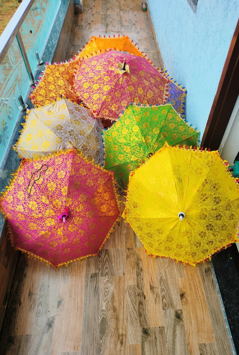 May include: A collection of colorful, decorative umbrellas with intricate floral patterns. The umbrellas are in shades of pink, yellow, green, and orange, with gold accents and tassel trim. The umbrellas are open and arranged on a wooden surface.