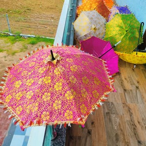 May include: A collection of colorful, decorative umbrellas. The umbrellas are in various colors, including pink, yellow, orange, green, and purple. The pink umbrella in the foreground has a gold floral pattern and decorative fringe.