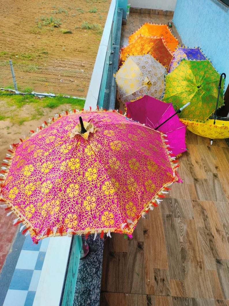May include: A collection of colorful, decorative umbrellas. The umbrellas are in various colors, including pink, yellow, orange, green, and purple. The pink umbrella in the foreground has a gold floral pattern and decorative fringe.