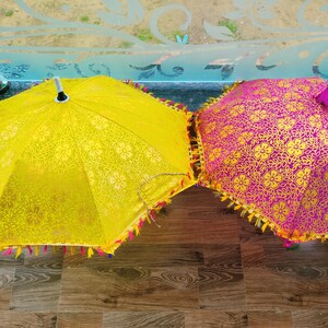 May include: Two decorative umbrellas. One is yellow with a floral pattern and a fringe trim. The other is magenta with a gold floral pattern and a fringe trim. The umbrellas are on a wooden surface.
