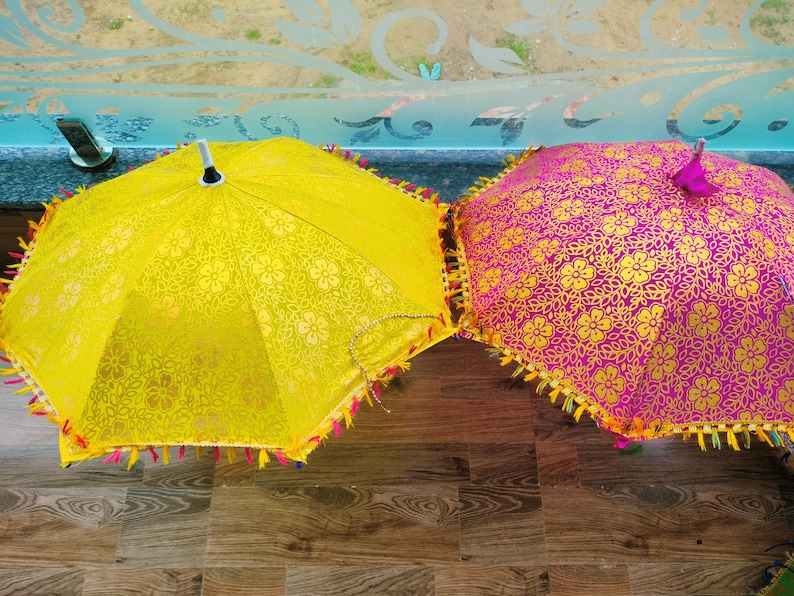 May include: Two decorative umbrellas. One is yellow with a floral pattern and a fringe trim. The other is magenta with a gold floral pattern and a fringe trim. The umbrellas are on a wooden surface.