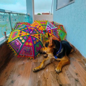 May include: A German Shepherd dog rests near several colorful, decorative umbrellas. The umbrellas feature intricate patterns in shades of pink, yellow, green, and blue, with decorative fringe. The dog has black and tan fur and a blue collar.