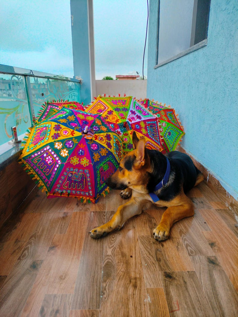 May include: A German Shepherd dog rests near several colorful, decorative umbrellas. The umbrellas feature intricate patterns in shades of pink, yellow, green, and blue, with decorative fringe. The dog has black and tan fur and a blue collar.