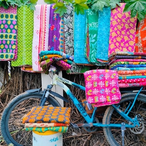 Op de afbeelding: Een blauwe fiets met kleurrijke textiel. Verschillende patronen dekens en stoffen in tinten roze, oranje, groen en blauw hangen boven de fiets, waardoor een levendige weergave van handgemaakte goederen ontstaat.