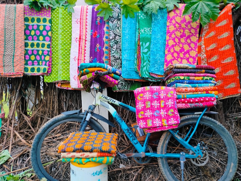 Op de afbeelding: Een blauwe fiets met kleurrijke textiel. Verschillende patronen dekens en stoffen in tinten roze, oranje, groen en blauw hangen boven de fiets, waardoor een levendige weergave van handgemaakte goederen ontstaat.
