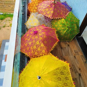 May include: A collection of colorful, open umbrellas with intricate floral patterns. The umbrellas are in shades of yellow, orange, pink, green, and purple, with decorative trim. The umbrellas are arranged on a wooden surface.