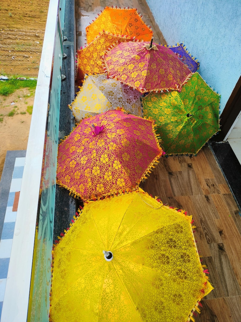May include: A collection of colorful, open umbrellas with intricate floral patterns. The umbrellas are in shades of yellow, orange, pink, green, and purple, with decorative trim. The umbrellas are arranged on a wooden surface.