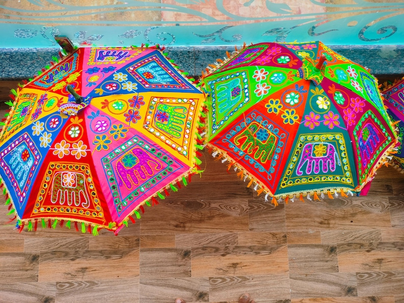 May include: Two colorful, open umbrellas with intricate embroidery and mirror work. The umbrellas feature vibrant patterns in red, yellow, blue, and green, with elephant and floral motifs. Each umbrella has a fringe trim.