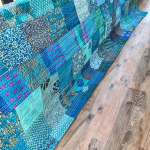 May include: A patchwork quilt in shades of blue, teal, and turquoise. The quilt is made of various fabric squares with different patterns, including floral and geometric designs. The quilt is draped over a surface, showcasing its texture and color variations.