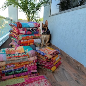 May include: Stacks of colorful patchwork quilts with various patterns and textures. The quilts are in shades of pink, orange, blue, and green. A German Shepherd dog sits near the quilts. The quilts are stacked on a wooden surface.