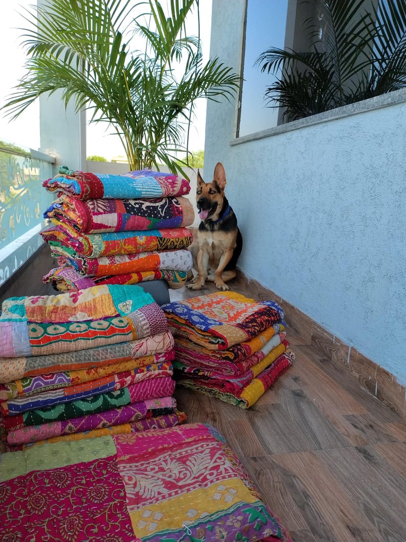 May include: Stacks of colorful patchwork quilts with various patterns and textures. The quilts are in shades of pink, orange, blue, and green. A German Shepherd dog sits near the quilts. The quilts are stacked on a wooden surface.