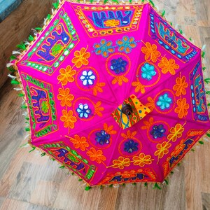 May include: A vibrant pink decorative umbrella with intricate embroidery and mirror accents. The umbrella features colorful floral and geometric patterns in orange, blue, and green. The edges are trimmed with green tassels.