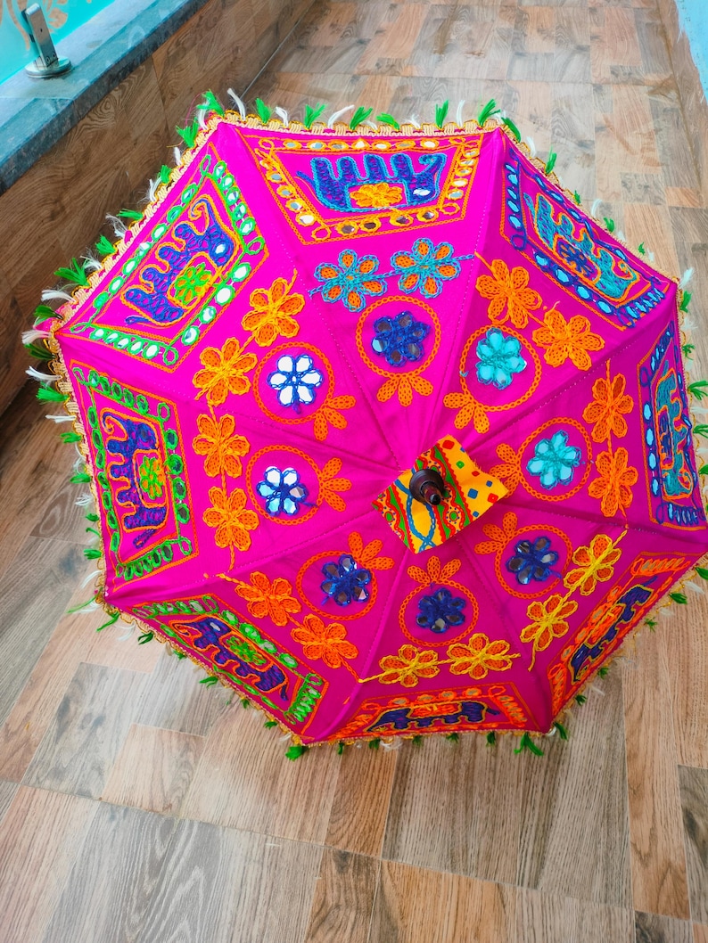 May include: A vibrant pink decorative umbrella with intricate embroidery and mirror accents. The umbrella features colorful floral and geometric patterns in orange, blue, and green. The edges are trimmed with green tassels.