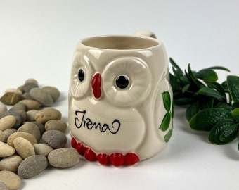 Może przedstawiać: Biały kubek ceramiczny w kształcie sowy z czerwonymi i czarnymi detalami. Kubek ma napisane na nim imię "Irena" czarnym, ozdobnym pismem.
