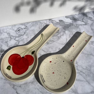 May include: Two ceramic spoon rests with a white background. One spoon rest has a red poppy flower design and the other has red hearts and black dots. Both spoon rests have the text "Rocca Verde" and "Handmade in Italy" on the bottom.