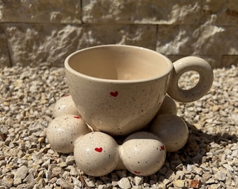 Może przedstawiać: Kubek ceramiczny w kolorze beżowym w białe kropki z czerwonym sercem na środku, stojący na beżowym ceramicznym podkładce w kształcie chmury.