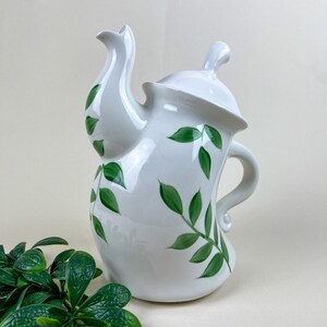May include: White ceramic teapot with a green leafy design. The teapot has a curved spout and a handle.
