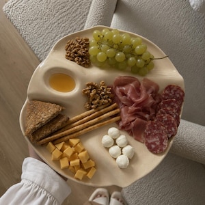 May include: A cream-coloured ceramic serving platter with an organic, irregular shape and a built-in circular dip bowl. The platter is filled with green grapes, walnuts, prosciutto, salami slices, mozzarella balls, cubed cheddar cheese, breadsticks, and toast.