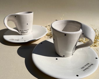 May include: Two white ceramic espresso cups with black polka dots and matching saucers. The saucers have the text "Maria Homar" and "Rosso d'oro" printed on them.