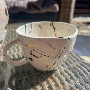 Könnte beinhalten: Weiße Keramik-Teetasse mit abstraktem Design in Schwarz und Gold. Die Tasse hat eine abgerundete Form und einen Henkel.