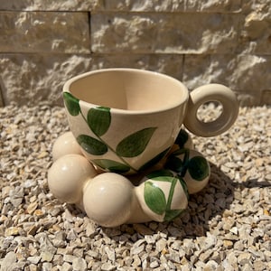 Puede incluir: Una taza de té de cerámica blanca con un diseño de hojas verdes se asienta sobre un posavasos de cerámica blanca con cuatro protuberancias redondas. El posavasos está decorado con hojas verdes.