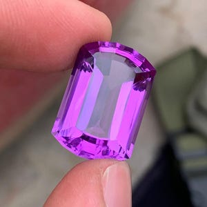 May include: A rectangular, faceted gemstone in a vivid purple shade. The gem is held between fingers, highlighting its polished surfaces and internal reflections. The cut of the stone is a rectangular shape with a curved top.