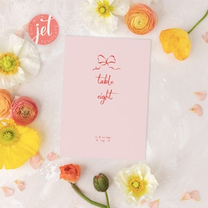 Könnte beinhalten: Eine rosa Tischkarten mit einer roten Schleife und dem Text "Tisch acht" in roter Schrift. Die Karte ist von gelben und rosa Blumen umgeben.