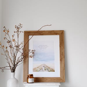 Könnte beinhalten: Ein gerahmter Druck mit hellem Holzrahmen, der eine Fotografie einer Person zeigt, die einen Weg entlanggeht. Eine weiße Vase mit getrockneten Zweigen und eine kleine Kerze stehen daneben auf einem weißen Tisch. Das Bild hat eine neutrale, beruhigende Ästhetik.