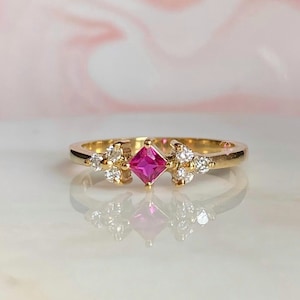May include: A gold ring with a pink square gemstone and smaller clear gemstones set around it.