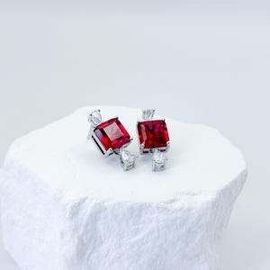 May include: A pair of silver-toned earrings featuring a central square-cut red gemstone. Each earring is accented with two small, clear, pear-shaped stones positioned at the top and bottom of the square setting, creating a symmetrical design.