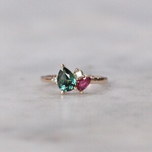 Puede incluir: Un anillo de oro rosa con una piedra preciosa verde en forma de pera, una piedra preciosa rosa más pequeña en forma de pera y una piedra preciosa blanca redonda.