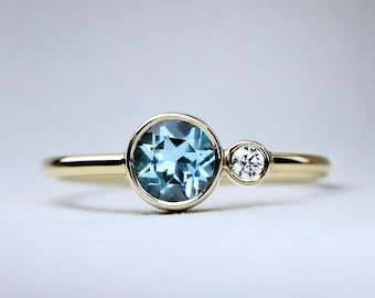 May include: A gold ring featuring a large, round, light blue gemstone and a smaller, clear, round gemstone. The gemstones are set in a bezel setting. The ring has a simple, elegant design.