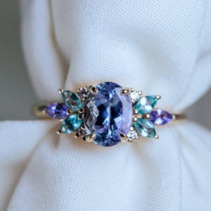 May include: A gold ring featuring a large, oval-cut, light purple gemstone as the centerpiece. The ring is adorned with smaller, teardrop-shaped gemstones in shades of blue and purple, and small white stones.