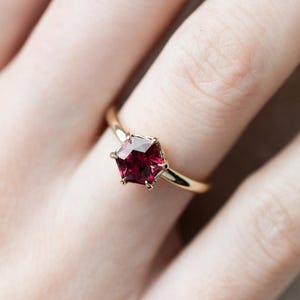 May include: A gold ring with a hexagonal, deep red gemstone. The gemstone is set in a gold bezel, and the band is plain gold. The ring is displayed on a finger, showcasing its elegant design and craftsmanship.