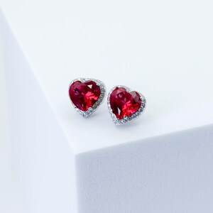 May include: A pair of heart-shaped stud earrings featuring vibrant red faceted stones set in silver-toned metal. Each earring is surrounded by a delicate halo of small, clear, sparkling stones that follow the heart-shaped outline of the center gem.