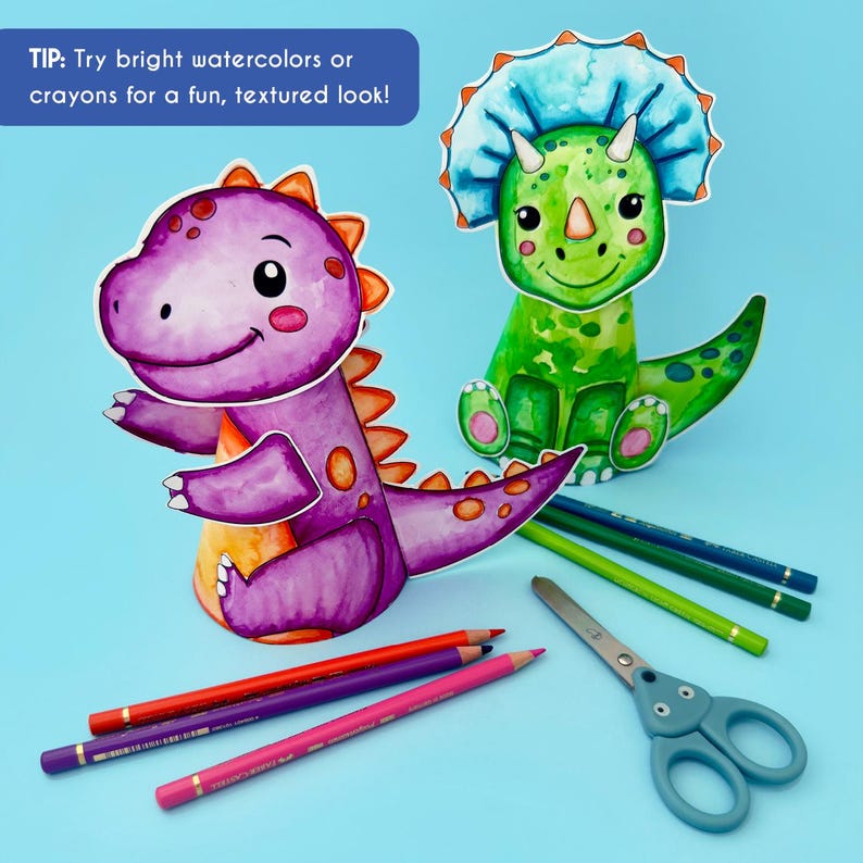 May include: Two colorful dinosaur cutouts, one purple and one green, sit on a blue surface. The purple dinosaur has orange accents, and the green one has a blue frill. Colored pencils and scissors are also present. Text reads: "Try bright watercolors or crayons for a fun, textured look!"