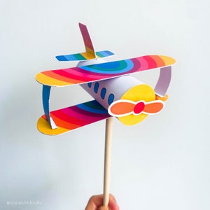 May include: A colorful paper airplane craft with a rainbow design and a yellow propeller. The airplane is attached to a wooden stick.