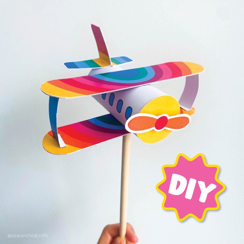 May include: A colorful DIY airplane craft made from a paper tube and colorful paper. The airplane has a rainbow design and a propeller. The craft is held by a hand and a pink starburst with the word "DIY" is in the background.