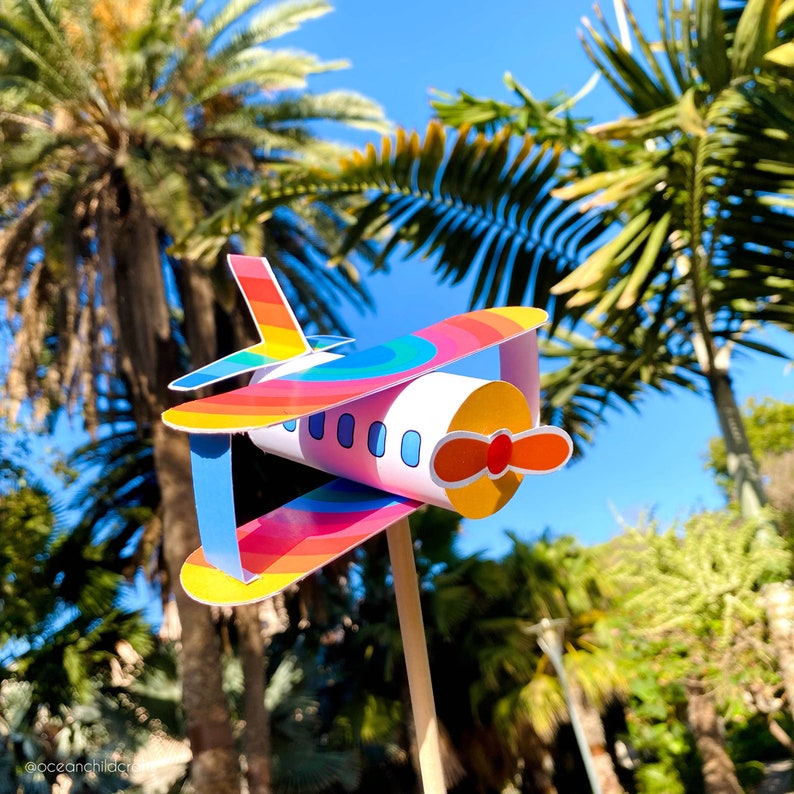 May include: A colorful paper airplane with a rainbow design and a white body. The airplane is attached to a wooden stick and is set against a background of palm trees and a blue sky.