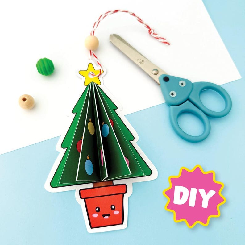 May include: A paper Christmas tree ornament with a green tree, decorated with colourful baubles, sits in a red pot. A yellow star tops the tree. Blue scissors and the word "DIY" are also in the image.