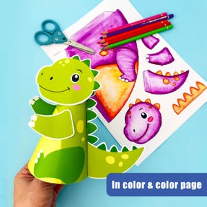 May include: A green and yellow dinosaur craft with a smiling face, held in a hand. The craft is next to a sheet with a purple dinosaur illustration, colored pencils, and scissors. The text "In color & color page" is visible.