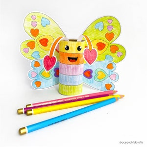 May include: A colorful butterfly craft made from a cardboard tube with a smiling face. The butterfly has wings with a heart pattern and is decorated with colored pencils.