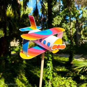 May include: A colorful paper airplane pinwheel with a rainbow design. The pinwheel is attached to a wooden stick and is set against a green background.