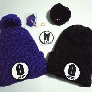 May include: Two knitted beanies, one purple and one black, each with a matching pom-pom. Both beanies feature a circular patch with a black and white graphic and the word "ARMY". Additional patches and craft supplies are also visible.