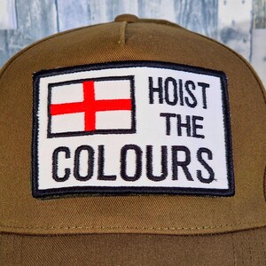 May include: A brown baseball cap with a rectangular patch displaying the English flag and the text "HOIST THE COLOURS" in black. The cap has a curved peak and a structured crown.