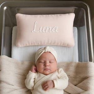Op de afbeelding: Een pasgeboren baby in een crèmekleurige geribbelde gebreide outfit en tulband ligt in een doorzichtige wieg. Een lichtroze kussen met de naam "Luna" in sierlijke letters geborduurd, ligt boven de baby. Een crèmekleurige gebreide deken bedekt de baby.