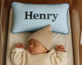 Puede incluir: Un bebé recién nacido con un gorro y un suéter de punto beige, acostado en una cuna. Una almohada azul claro con el nombre "Henry" en letras negras está detrás del bebé. El bebé lleva un pañal y calcetines grises.