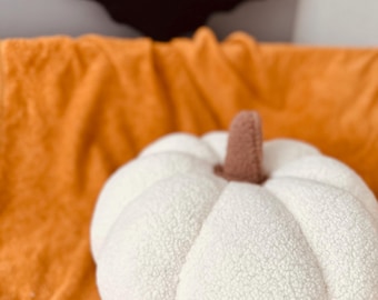 May include: A white, plush pumpkin with a brown stem sits on an orange surface. A black bat decoration is in the background. The pumpkin is a soft, textured decorative item, perfect for fall or Halloween.