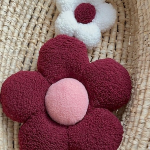 May include: Two flower-shaped decorative pillows. One is white with a burgundy center, and the other is burgundy with a pink center. The pillows have a soft, textured appearance and are placed inside a woven basket.